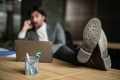 Close up of a businessman's shoes on the table in the office while he is talking on cell phone and using laptop. Copy space.