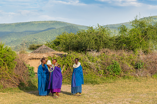TANZANIA, AFRICA-FEBRUARY 15, 2020: Four Massai Women and one Baby with traditional Dresses stay smiling at Sunset together. In the Background a Part of a Hut, Plants and Mountain Landscape