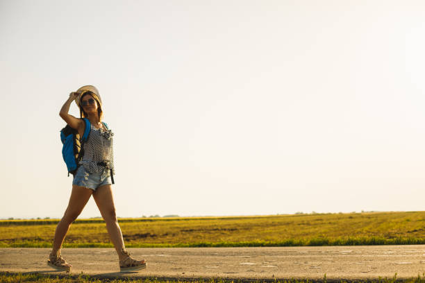 Wide shot of rural road where one adventurous joyful woman is backpacking to explore natural destinations.
