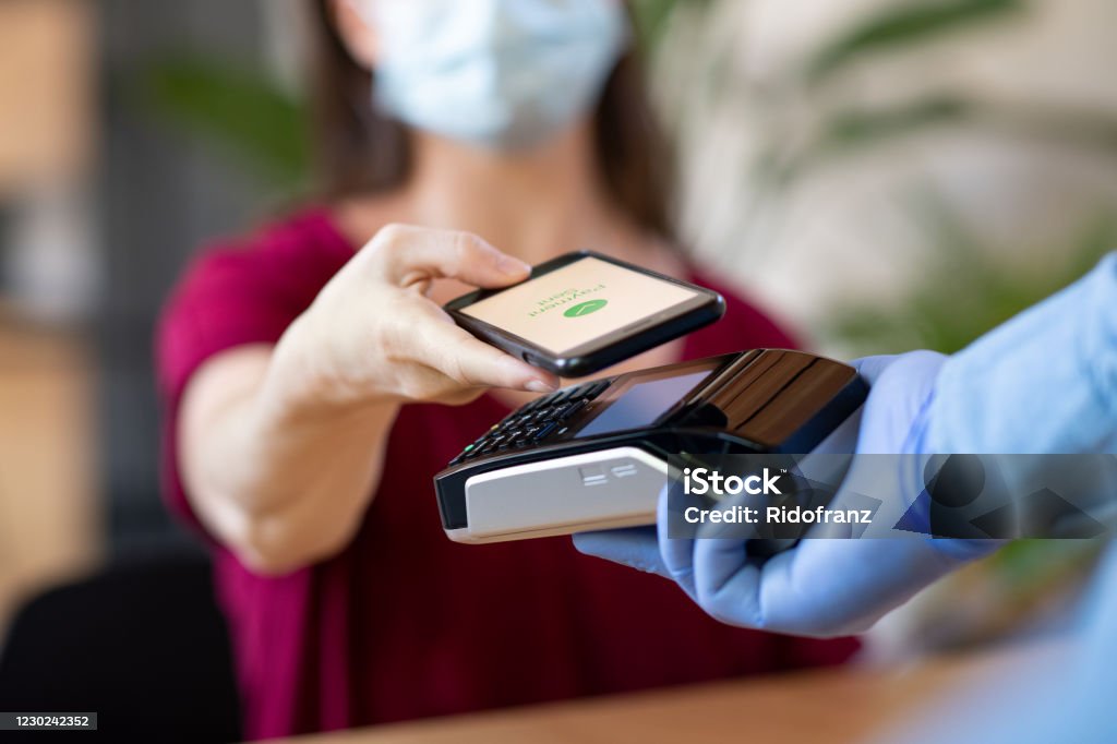 Contactless smartphone payment Close up hand of customer paying with smartphone. Cashier hand holding credit card reader machine and wearing protective disposable gloves at bar counter, while client holding phone for NFC payment. Woman wearing face mask while paying bill with mobile phone during Covid-19 pandemic. Paying Stock Photo
