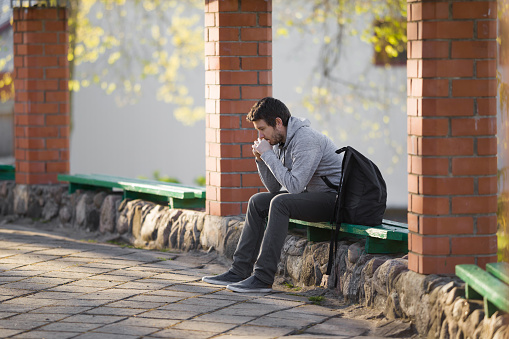 One young sad adult man sitting alone on wooden bench. Thinking about life. Side view.