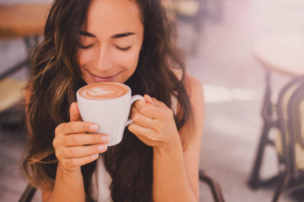 Young beautiful happy woman enjoying cappuccino in a cafe Young beautiful happy woman with long curly hair enjoying cappuccino in a street cafe hot chocolate photos stock pictures, royalty-free photos & images