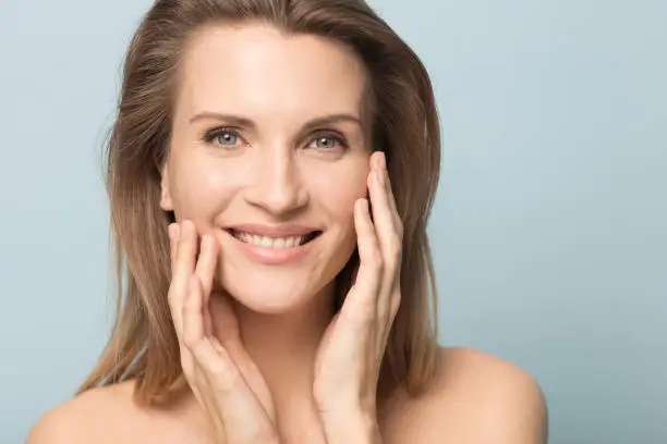 Head shot portrait smiling woman touching perfect smooth face skin, looking at camera, attractive young female with bare shoulders, natural beauty concept, close up isolated on studio background