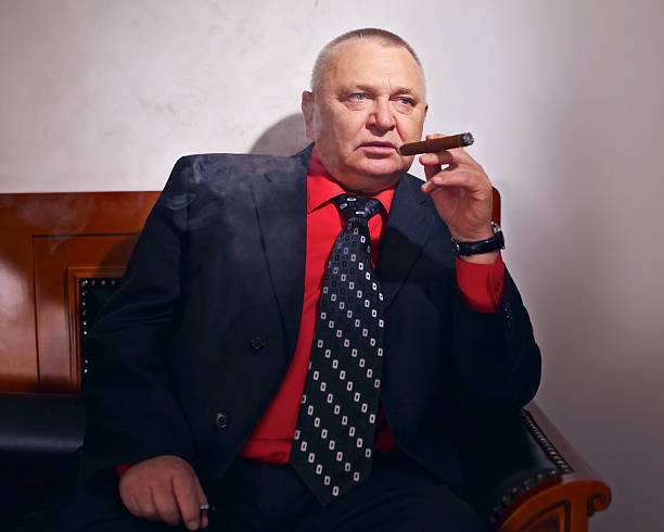 Big boss smoking cigar Middle aged businessman wearing red shirt, black coat and tie sitting on leather sofa and smoking cigar indoor mafia boss stock pictures, royalty-free photos & images