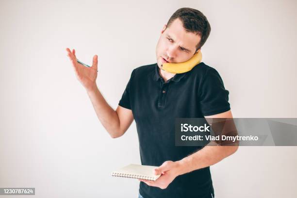Young Man Isolated Over White Background Busy Serious Guy Using Banana As A Smartphone And Talking On It Hold Notebook In Hand And Explaining Casual Businessman Stock Photo - Download Image Now