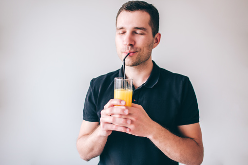 Young man isolated over background. Delightful happy guy drinking orange juice through plastic straw. Hold glass in hands. Positive cheerful man