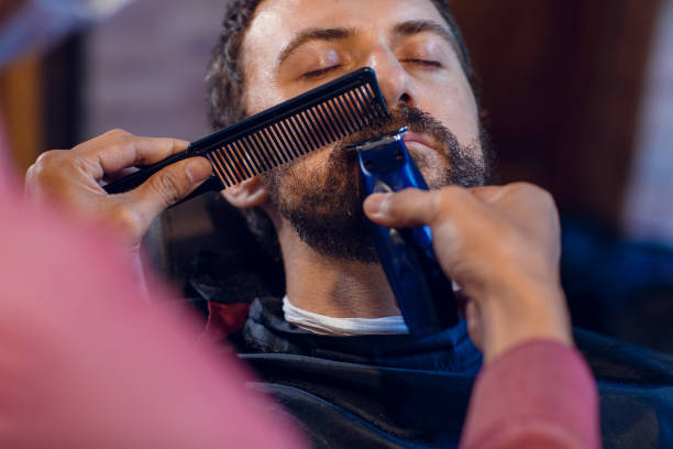 istock Beard styling. Barber shaping beard of handsome man using a trimmer. Skilled barber trimming beard to his client 1230844235