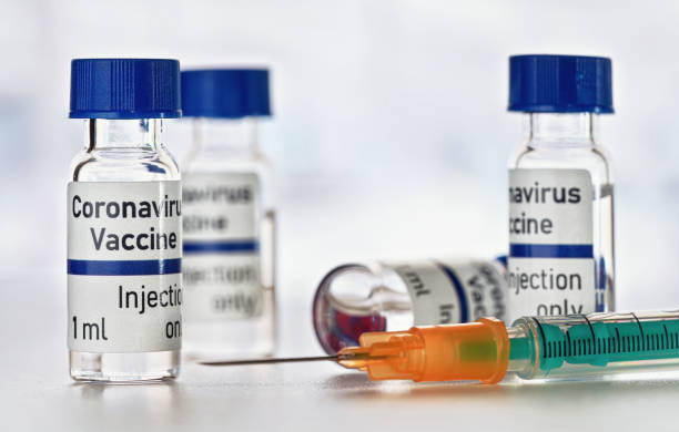 istock Coronavirus Covid 19 vaccine concept (own design, not real product) - small glass vials with blue caps on white table, green orange syringe near, closeup detail 1230853349
