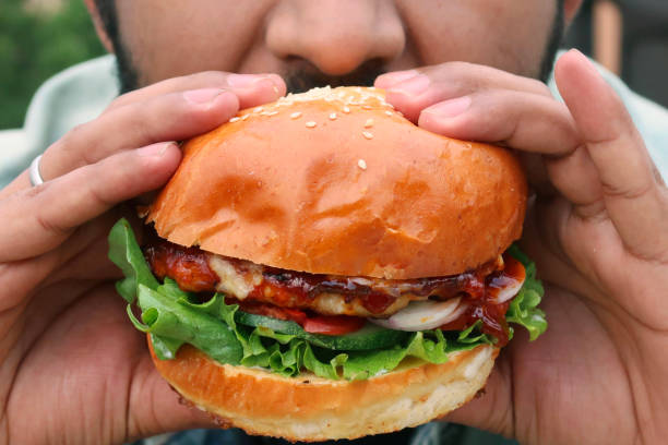 Image of unrecognizable man taking a bite of sesame seed bun chicken burger with chicken patty, onion rings, tomato slices, cucumber and lettuce leaves and  tomato ketchup sauce in hands