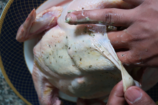 Stock photo of unrecognised man rubbing the butter and herbs under the skin of whole raw chicken to make roast dinner in kitchen worktop in non stick baking tray and ready to cook, Sunday roast dinner with crispy skin, herb stuffing in chicken