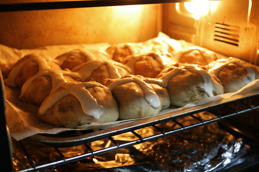 Stock photo showing homemade Easter hot cross buns in hot oven in greaseproof paper lined baking oven tray, home baking concept.\nThese traditional spiced sweet buns are made and sold over the Easter period, with the cross symbol on the glazed top being made from a flour and water paste, and symbolising Christianity.