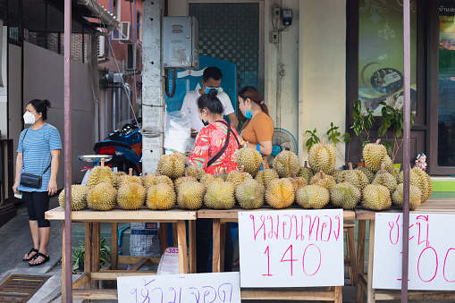 Durian street market stall and thai people in Bangkok during covid-19 lockdown in Ladprao. On table are many durian fruits. In scene are some people and women behind table. A woman is standing at left side. All are having masks, one has mask only around neck and throat.
