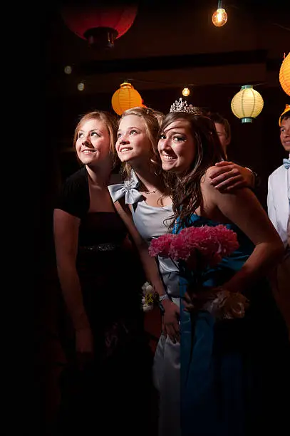A group of teenagers at a formal dance smile for a picture. One teenage girl stops to look away at the camera.