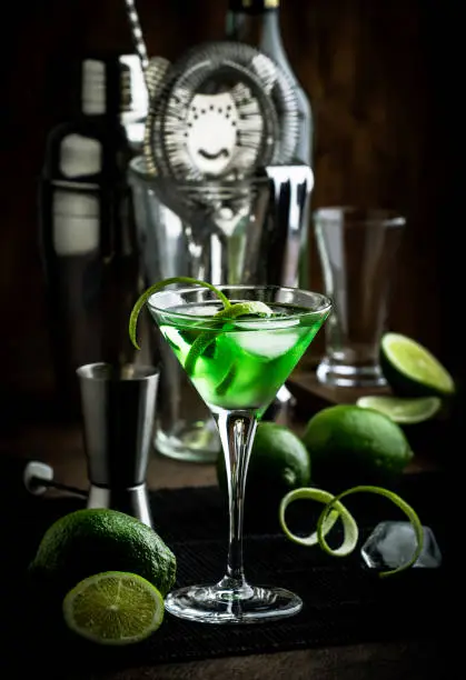 Green martini alcoholic cocktail in glass with dry gin, vermouth, liquor, lime zest and ice, steel bar tools, dark background