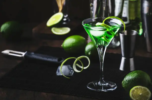 Green martini alcoholic cocktail in glass with dry gin, vermouth, liquor, lime zest and ice, steel bar tools, dark background