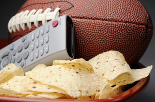 Stock photo of NFL style football with TV remote control and bowl of tortilla chips. 