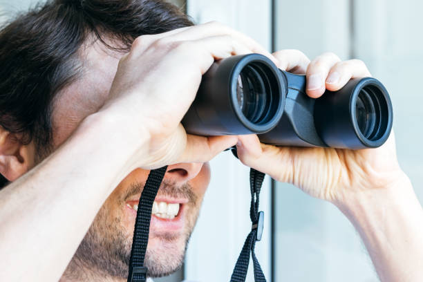 istock Handsome smiling bearded man looking through binoculars at the window with a view of the neighboring house for observing neighbors, environment or nature during quarantine. Selective focus. Closeup 1231376321
