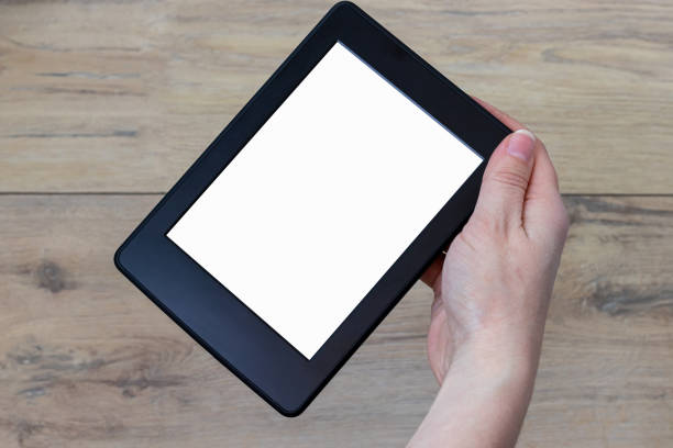 istock A modern black electronic book with a white empty blank screen rotated 45 degrees in female hand against a blurred wooden background. Mockup tablet template closeup 1231385576