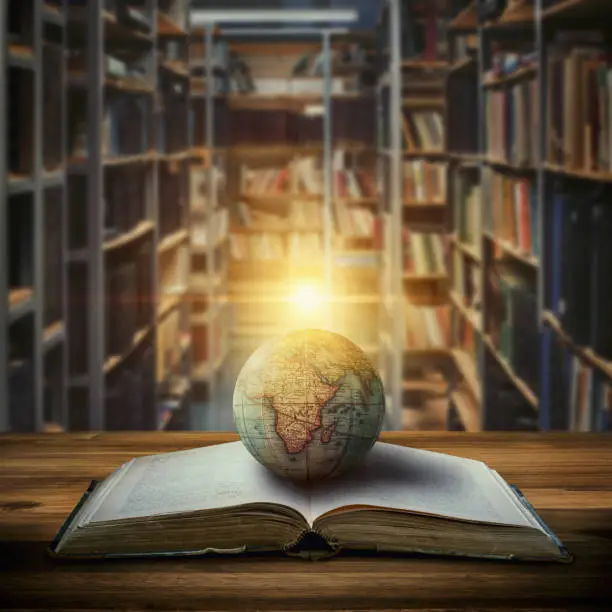 Photo of An old globe lying on an open book against the background of bookshelves in a library. Selective focus. Retro style.