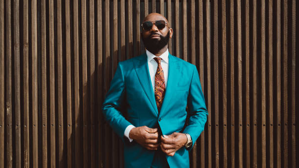 A handsome black guy in a blue suit A handsome mature bald bearded African man in a sunglasses and a fashionable blue or teal costume with a tie is standing in front of a wall made of striped wooden timbers and fastening a suit button necktie photos stock pictures, royalty-free photos & images