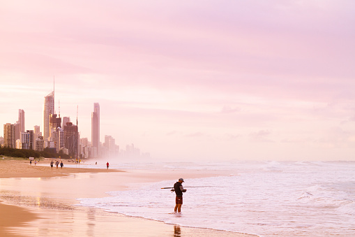 April 29, 2018 - Broadbeach - Mermaid Waters, Gold Coast, Queensland, Australia: Young man flyfishing at the sea with Gold Coast Skyline at sunset on back.