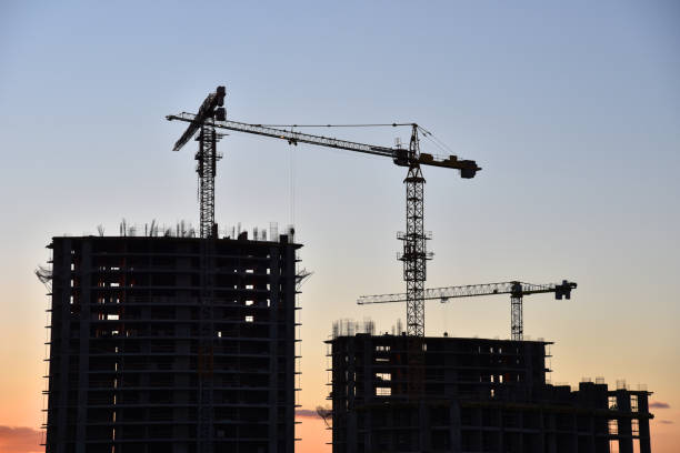 istock Silhouettes of tower cranes constructing a new residential building at a construction site on sunset background. Crane lifting a concrete bucket. Builders pouring cement mix for formworks. Soft focus 1231673540