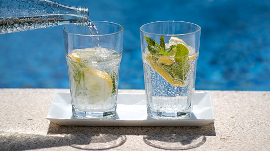 Mineral water from a bottle is poured into a glass with lemon and mint. Pouring sparkling water with bubbles in sunny day on blurred pool or sea background. Refreshing beverages by the pool.