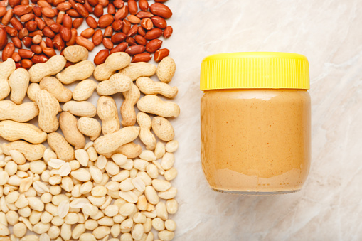 Creamy peanut paste in glass jar with yellow cap and peanuts in the shell and peeled peanuts. Creamy peanut paste flat lay with place for text on white marble background for cooking breakfast