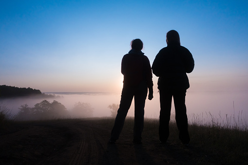 silhouette of two peple looking at sunrise on foggy valley, standing together ot the hill