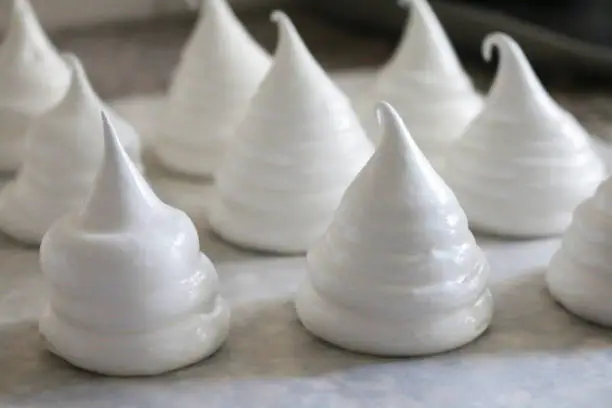 Stock photo showing a baking tray lined with wax paper containing rows of piped raw meringue peaks.