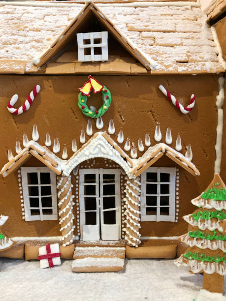 Close-up image of homemade Christmas gingerbread biscuit house decorated with icing and candy sweets on kitchen worktop with Christmas trees