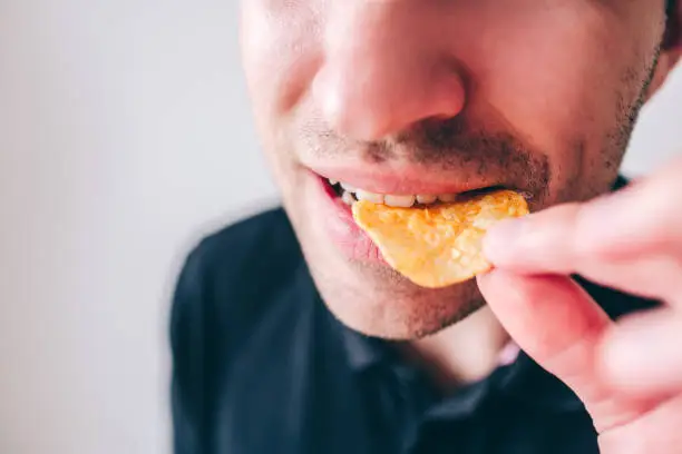 Photo of Young man isolated over background. Cut view and close up of guy biting piece of potato chips with teeth. Holding in it hand. Tasty delicious fast food.