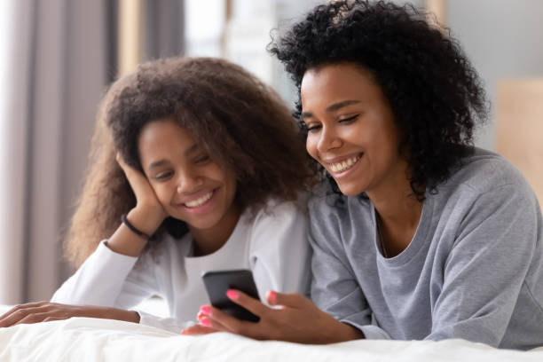 mère et descendant africains heureux utilisant le smartphone se trouvant sur le bâti - teenager african descent laughing adolescence photos et images de collection