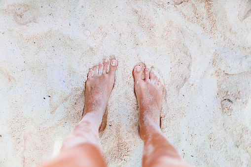 A woman's feet in black flip flops on the white sands of the beach as the waves roll in.