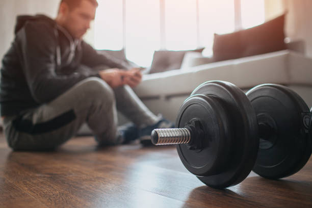 istock Young ordinary man go in for sport at home. Cut view with blurred background where workout beginner sit on floor and has rest after hard workout. Big dumbbells on floor. Sport amateur. 1232347021