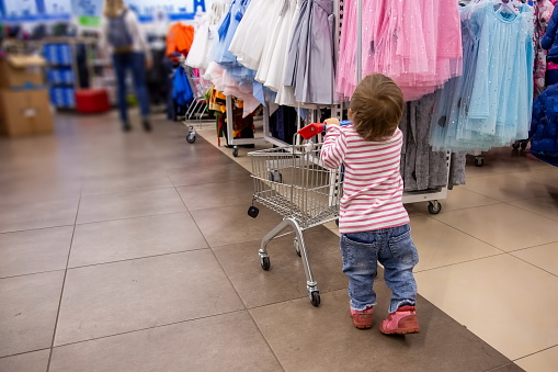 black friday shopping with children. a little cute toddler in jeans and a striped pink sweater is standing with a shopping cart in front of hangers with clothes. close-up, soft focus, blur background