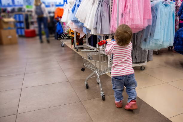 black friday shopping with children. a little cute toddler in jeans and a striped pink sweater is standing with a shopping cart in front of hangers with clothes. close-up, soft focus, blur background stock photo