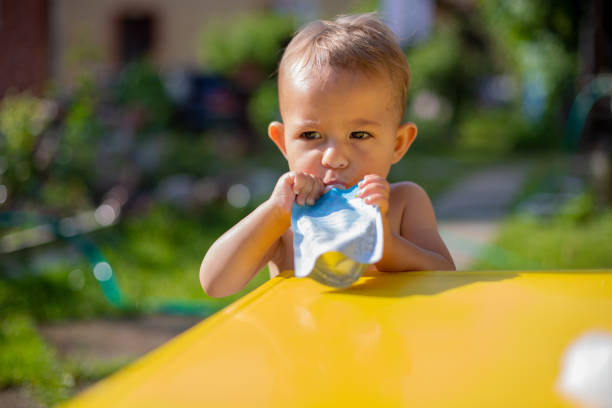 cute little caucasian baby eating fruit puree in pouch and looking into the camera in front of the yellow table. close up, on the background is  a green garden on a sunny day in blur stock photo