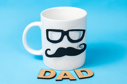 White mug with glasses and mustache and the word dad made from wooden letters on blue background