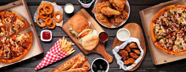 Table scene of assorted take out or delivery foods, top down view on a dark wood banner Table scene of assorted take out or delivery foods. Hamburgers, pizza, fried chicken and sides. Top down view on a dark wood banner background. fast food stock pictures, royalty-free photos & images