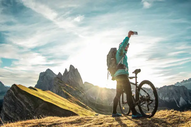 Young man with mountain bike taking selfies on Seceda mountain peak at sunrise. Puez Odle, Trentino, Dolomites, Italy.