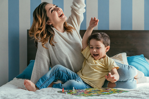 excited mother and son showing winner gestures while playing board game on bed