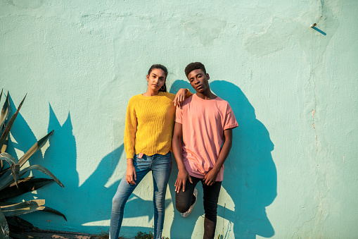 Portrait of Young couple. They are looking at camera while leaning on light blue wall.