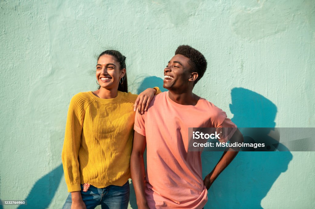 Portrait of two smiling couple looking away. Portrait of Young couple. They are looking away laughing and leaning on light blue wall Friendship Stock Photo