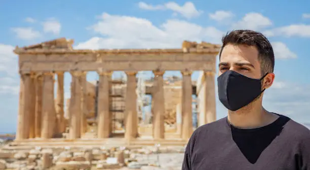 Photo of Athens Acropolis, Greece coronavirus days. Young man wearing protective face mask on Parthenon temple and blue sky background.