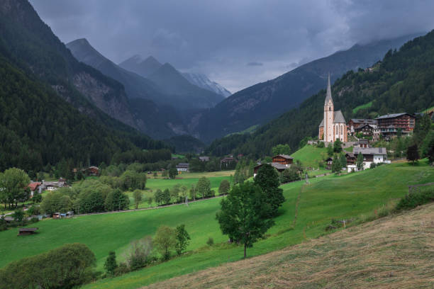 Mountain village Heiligenblut with picturesque church St. Vinzenz and green fields, rain clouds in the sky, Carinthia Austria