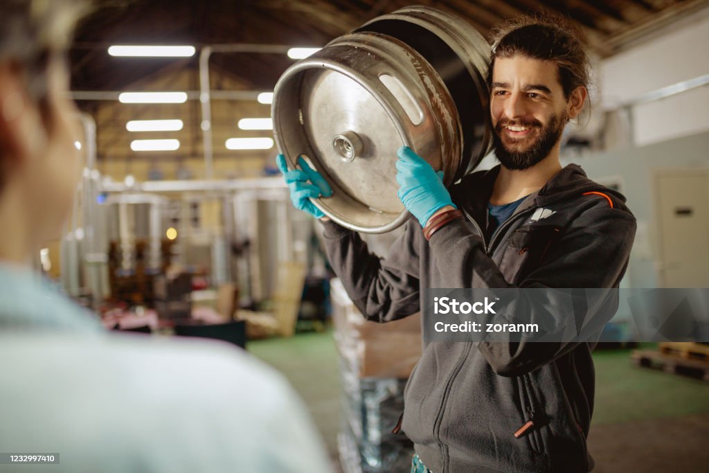 Worker carrying beer barrel on shoulder and smiling at manager Smiling worker carrying beer keg out of the warehouse for distribution and talking to female manager Keg Stock Photo