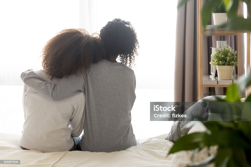 Loving african single mother embrace teen daughter sit on bed Loving african american single mother sister embrace teen daughter sit on bed looking at window, parent mom hug support protect teenage girl, family trust hope talk understanding concept, rear view Mental Health Stock Photo