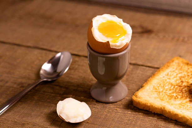 abriu ovo cozido em uma xícara de ovo com gema com uma fatia de pão torrado e uma colher em um fundo de madeira marrom natural. - sandwich breakfast boiled egg close up - fotografias e filmes do acervo