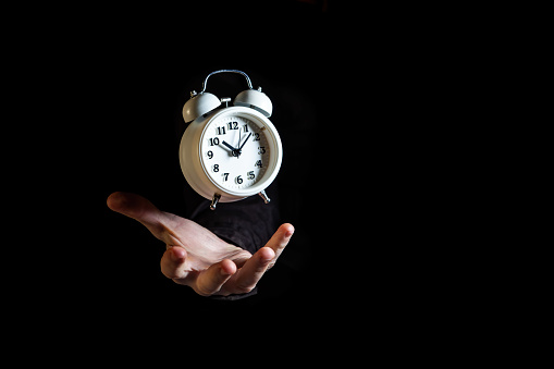White alarm clock suspended in the air above white male hand on black background with copy space - time concept